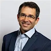 A smiling man in a suit with glasses, standing against a plain background.