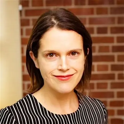 A woman with shoulder-length hair wearing a striped black and white shirt, smiling in front of a brick wall.